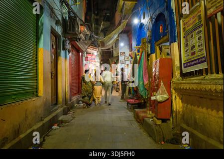 Varanasi, Inde - 17 mars 2024 : plusieurs personnes marchent dans une ruelle étroite faiblement éclairée avec des volets fermés, des textiles suspendus, des panneaux muraux en hindi et des déchets éparpillés visibles la nuit. Banque D'Images