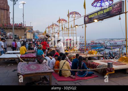 Varanasi, Inde - 17 mars 2024 : des groupes de personnes sont assis sur des plates-formes et des tapis à Dashashwamedh Ghat, avec des parapluies décorés, des objets rituels et une pancarte indiquant « Gangotri Seva Samiti » visible, alors que les foules et les bateaux se rassemblent le long du Gange lors d'une cérémonie en soirée. Banque D'Images