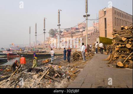 Varanasi, Inde - 19 mars 2024 : des gens sont vus marcher, se rassembler et travailler près de piles de bois et de restes de crémation à Manikarnika Ghat le long du Gange, avec des bateaux amarrés au bord de l'eau et des bâtiments historiques et de grands lampadaires visibles en arrière-plan pendant la journée. Banque D'Images