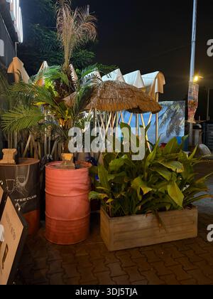 Perak, Malaisie - 24 janvier 2026. Un coin salon extérieur nocturne avec des plantes tropicales, un parapluie de chaume et des barils d'huile industriels utilisés comme tables Banque D'Images