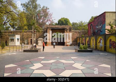 Prayagraj, Inde - 21 mars 2024 : plusieurs personnes sont vues marcher et se tenir debout près de la porte d'entrée nord de Khusro Bagh, avec un mur de pierre, des cabines de sécurité, des palmiers et des peintures murales colorées sur les murs environnants pendant la journée. Banque D'Images