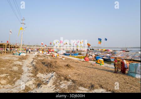 Prayagraj, Inde - 21 mars 2024 : de nombreux bateaux couverts et découverts sont amarrés le long de la rive sablonneuse de Sangam, avec des drapeaux colorés, des structures temporaires, des gens en vêtements traditionnels et des poteaux utilitaires visibles sous un ciel dégagé. Banque D'Images