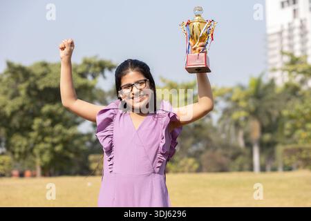 Heureux enfant de fille indienne tenant le trophée en plein air dans le parc célébrant la victoire. Réussite et succès. Banque D'Images