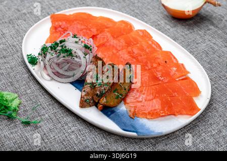 Gros plan d'un plateau vibrant de truites fumées ou séchées tranchées (gravlax), servies avec des rondelles d'oignon rouge finement tranchées, des quartiers de pommes de terre rôties aux herbes, et Banque D'Images