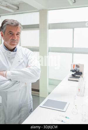 Homme mature debout à gauche de la paillasse portant une blouse de laboratoire, une tablette, un microscope, des tubes à essai, un espace de copie. Scientifique, laboratoire, recherche, concentration, précision Banque D'Images