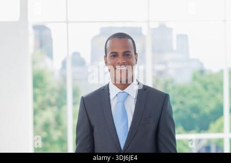 Homme afro-américain debout et souriant dans le bureau moderne portant costume et cravate devant les fenêtres de grille. Portrait, professionnel, exécutif, entreprise, WOR Banque D'Images