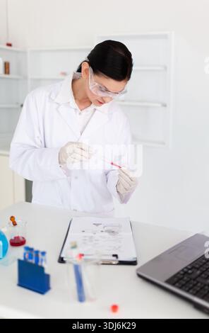 Le tube à essai transparent contenant du liquide rouge repose sur un banc de laboratoire blanc près de l'ordinateur portable. Scientifique, laboratoire, lunettes, gants, presse-papiers, pipette, life esc Banque D'Images