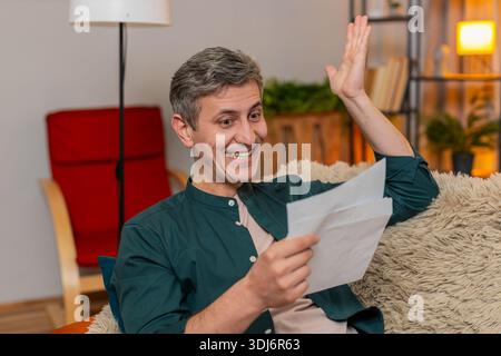 Jeune homme heureux à la maison ouvre la lettre et célèbre joyeusement les grandes nouvelles, levant le poing avec joie. Caucasien Guy sur le canapé se sent excité par l'offre d'emploi, l'approbation de prêt ou le résultat réussi de l'examen scolaire. Banque D'Images