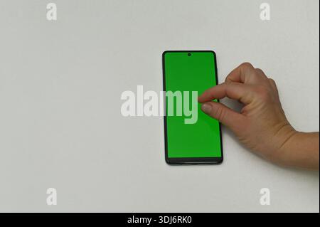 la main humaine tape sur l'écran vert d'un smartphone positionné verticalement sur un fond blanc. téléphone portable avec une zone chromée verte est utilisé avec Banque D'Images