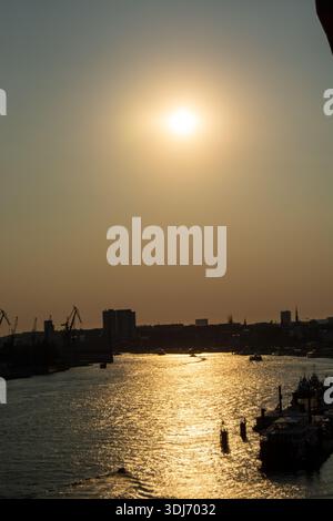 Un coucher de soleil serein sur une rivière, avec le soleil se reflétant sur l'eau, silhouettes de grues et de bâtiments en arrière-plan Banque D'Images