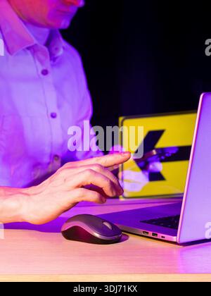Le doigt vise l'icône de flèche gauche-bas jaune sur la feuille claire positionnée à côté de l'écran d'ordinateur portable dans un environnement coloré de néon. Banque D'Images
