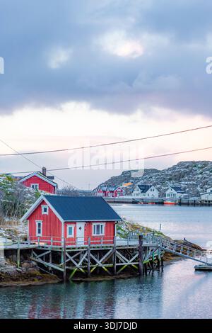 Petite maison rouge se trouve sur une jetée en bois surplombant un plan d'eau. Le ciel est nuageux et le soleil se couche, créant une atmosphère de mauvaise humeur Banque D'Images