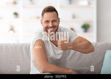 Un gars heureux approuvant les vaccinations, montrant du plâtre médical sur l'épaule après l'injection et en faisant des gestes de pouce vers le haut, espace libre Banque D'Images