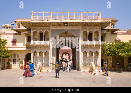Jaipur, Inde - 16 mars 2022 : la porte d'entrée principale du City Palace est représentée avec des sculptures en pierre blanche complexes, des balcons, deux statues d'éléphants en pierre flanquant la porte, plusieurs personnes marchant à travers l'arche, et des drapeaux visibles sur le toit sous un ciel bleu clair. Banque D'Images