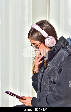 Jeune femme / adolescente portant des écouteurs, fumant et tenant un smartphone - Loches, Indre-et-Loire (37), France. Banque D'Images
