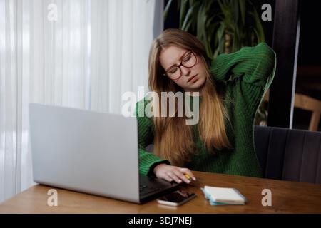 Jeune femme dans des lunettes tenant le cou dans la douleur, éprouvant l'inconfort de longues heures de travail à l'ordinateur portable. Banque D'Images