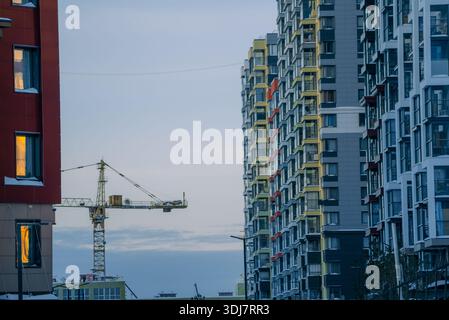 Grue de construction opérant entre des immeubles d'appartements modernes reflétant la lumière du coucher du soleil sur les fenêtres, symbolisant le développement urbain et la croissance dans un ciel urbain changeant Banque D'Images