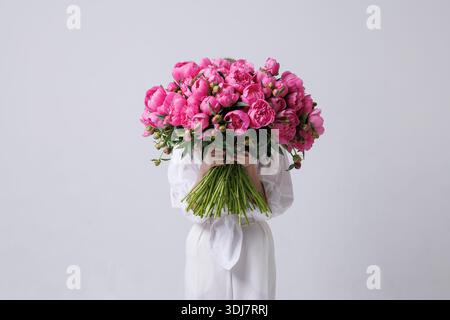 Femme élégante avec la tête couverte de pivoines roses posant dans une tenue blanche sur un fond minimal, concept de mode florale artistique. Banque D'Images