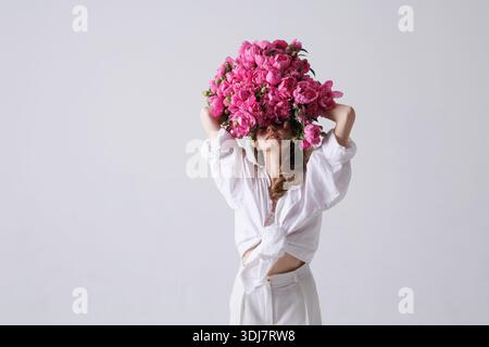 Femme élégante avec la tête couverte de pivoines roses posant dans une tenue blanche sur un fond minimal, concept de mode florale artistique. Banque D'Images