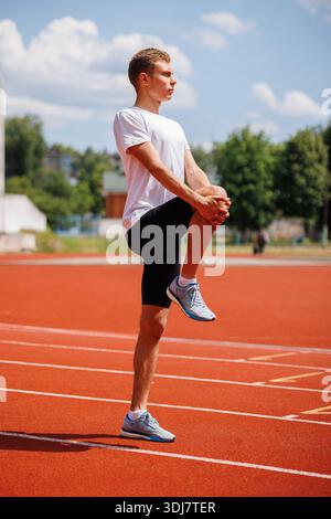 Homme athlétique qui s'étire avant de courir sur une piste en plein air, qui effectue un échauffement debout du genou à la poitrine par une journée ensoleillée. Banque D'Images