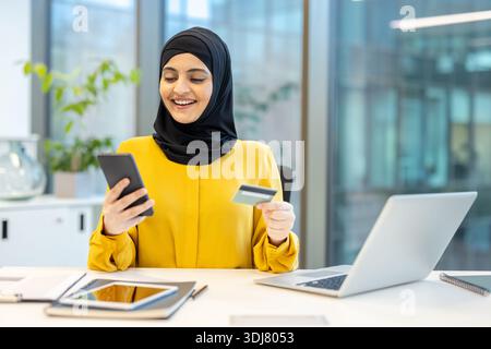 Femme d'affaires musulmane portant un hijab assise à un bureau, souriant tout en tenant un smartphone et une carte de crédit, en utilisant les services bancaires en ligne pour effectuer une transaction de commerce électronique Banque D'Images
