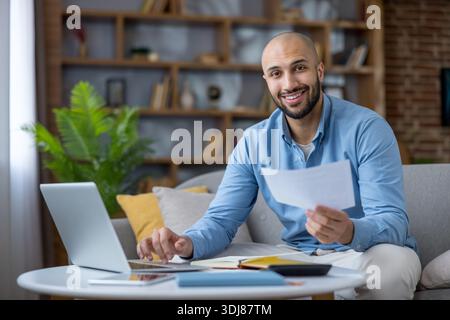 Jeune homme souriant à la caméra tout en travaillant sur un ordinateur portable, tenant des documents, calculant le budget et gérant les finances à partir de la configuration du bureau à domicile dans un salon confortable Banque D'Images