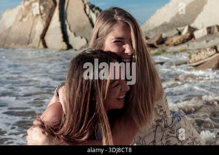 Deux amies joyeuses appréciant une balade en piggyback par les vagues de l'océan et les rochers Banque D'Images