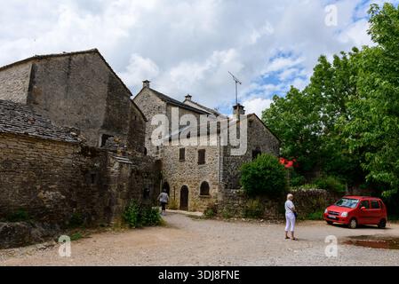 La Couvertoirade, France - 26 juin 2022 : plusieurs bâtiments historiques en pierre avec des toits d'ardoise, une voiture rouge garée sur une zone de gravier, et deux personnes marchant et debout dans le village médiéval sous un ciel partiellement nuageux. Banque D'Images