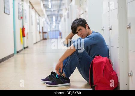 Étudiant masculin assis sur le sol à côté de casiers blancs dans le couloir de l'école avec sac à dos rouge, espace de copie. Contemplatif, couloir, intérieur, denim, sarcelle, pu Banque D'Images
