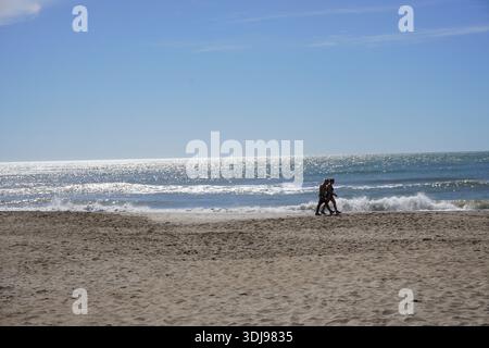Les silhouettes d'un homme et d'une femme marchant le long d'une large plage de sable fin à côté de l'océan étincelant par une journée claire et ensoleillée. Banque D'Images