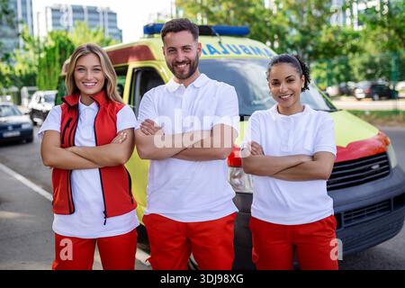 Groupe multiethnique de paramédicaux debout à côté d'une ambulance avec portes ouvertes. Leur collègue portant un sac médical pour traumatismes. Ils sourient Banque D'Images