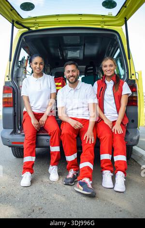 Groupe multiethnique de paramédicaux debout à côté d'une ambulance avec portes ouvertes. Leur collègue portant un sac médical pour traumatismes. Ils sourient Banque D'Images
