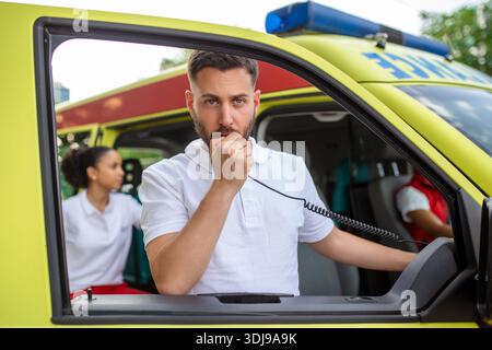 Un adulte beau paramédical masculin parle sur une radio portable alors qu'il est assis dans une ambulance à l'extérieur d'une clinique. Femme afro-américaine paramédicale stan Banque D'Images