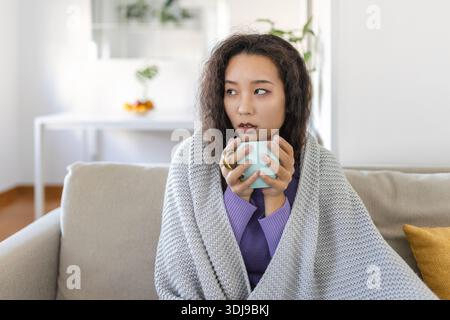 Malade jeune femme asiatique assise à la maison dans le lit avec une tasse de thé chaud et mouchoir. Rhumes saisonniers, toux, écoulement nasal, infections virales, traiteurs à domicile Banque D'Images