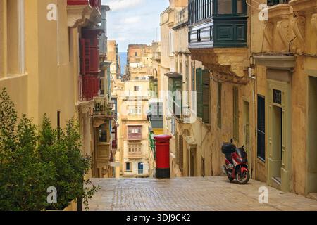 Rue maltaise avec balcons traditionnels en bois, une boîte aux lettres rouge vintage et une vue lointaine sur la mer Méditerranée. Banque D'Images