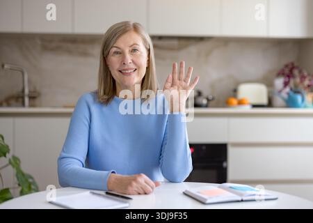 Femme mature souriante assise à une table dans sa cuisine, agitant sa main tout en faisant un appel vidéo, représentant la communication à distance et la connexion amicale Banque D'Images