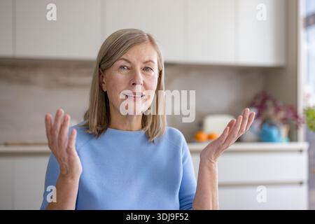 Femme senior communiquant avec des gestes de la main et une expression amicale lors d'une réunion virtuelle ou d'une conversation en ligne, en restant connectée depuis sa cuisine moderne Banque D'Images