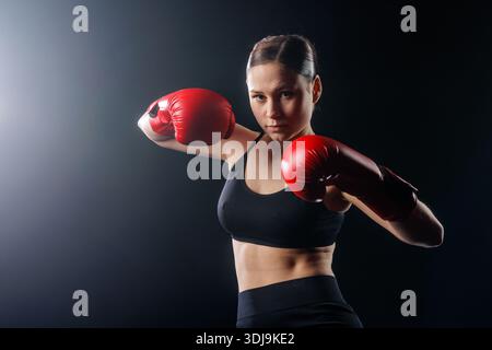 Jeune boxeur féminin dans des gants rouges dans la position de combat sur fond de studio sombre avec espace de copie pour la bannière de site Web ou la publicité sociale. Banque D'Images