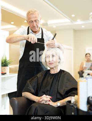 Le maître expérimenté de l'homme senior fait effectue la coupe de cheveux pour la femme mature avec des ciseaux Banque D'Images