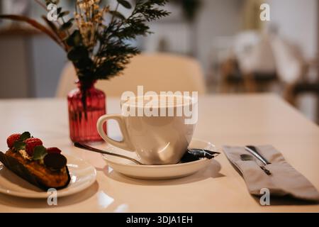 Une scène de café minimaliste avec une tasse de cappuccino servi sur une soucoupe blanche, accompagné d'un éclair garni de crème, fraises fraîches. Un petit rose Banque D'Images