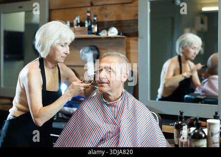 Le maître expérimenté de la femme mature fait effectue la coupe de cheveux pour l'homme âgé avec tondeuse Banque D'Images