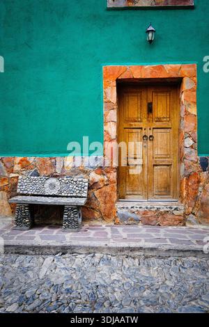 Une porte en bois accueillante avec un banc en pierre devant un mur vert vibrant, ajoutant du charme au paysage architectural Banque D'Images