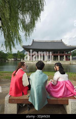 Séoul, Corée du Sud : trois visiteurs dans un hanbok traditionnel coloré assis près de l'étang au palais de Gyeongbokgung avec pavillon historique en arrière-plan Banque D'Images