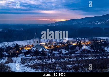 Un village d'hiver tranquille à l'aube, avec une église blanche entourée de toits enneigés et de lampadaires chauds, avec des collines lointaines Banque D'Images