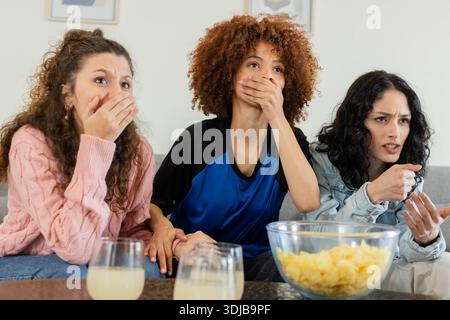 Diverses amies féminines haletant, faisant des gestes et se penchant dans le salon avec des frites et un pull rose Banque D'Images