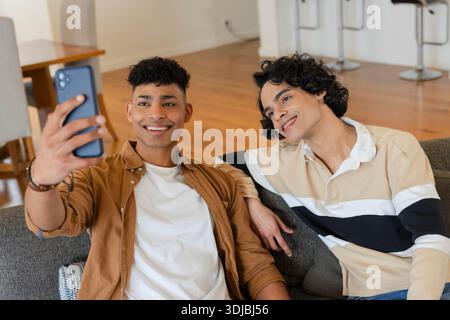Deux amis masculins tenant un smartphone prenant selfie tout en étant assis sur un canapé gris dans un appartement moderne Banque D'Images