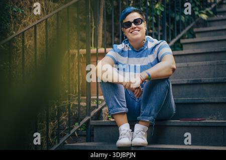 Une personne non binaire souriante avec des cheveux bleus courts et des lunettes de soleil est assise sur des escaliers en pierre extérieurs, représentant l'identité de genre authentique et le visibi LGBTQ+ Banque D'Images