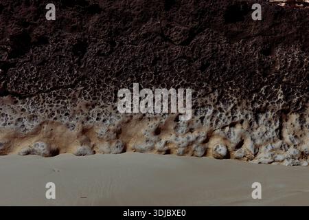 Une vue aérienne capture le contraste spectaculaire entre les formations rocheuses volcaniques poreuses et le sable beige lisse sur une plage de la péninsule arabique. L'obscurité Banque D'Images