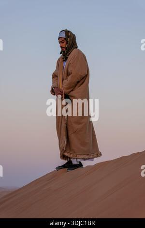 Un homme omanais en tenue traditionnelle se tient au sommet d'une dune du désert, baigné dans la douce lueur du crépuscule. Cela évoque un sentiment de voyage culturel et serein a Banque D'Images