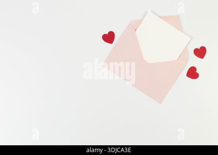 Vue de dessus de l'enveloppe rose, carte blanche sur fond blanc. Saint Valentin, cœurs rouges, amour. Post pose à plat, espace de copie. Banque D'Images